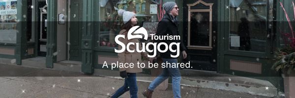 couple walking in downtown Port Perry with light snowfall and overlayed banner housing Scugog Tourism logo and text reading 'A place to be shared.'